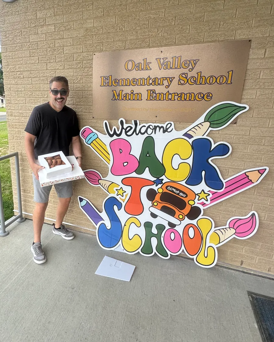 Eric holding a box of treats next to a Welcome Back to School sign at Oak Valley Elementary School