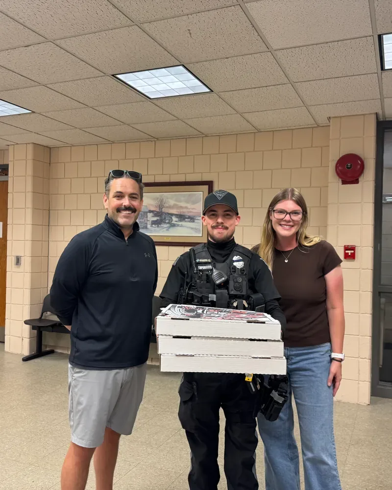 Eric and a teammate with a police officer holding pizza boxes inside a police station